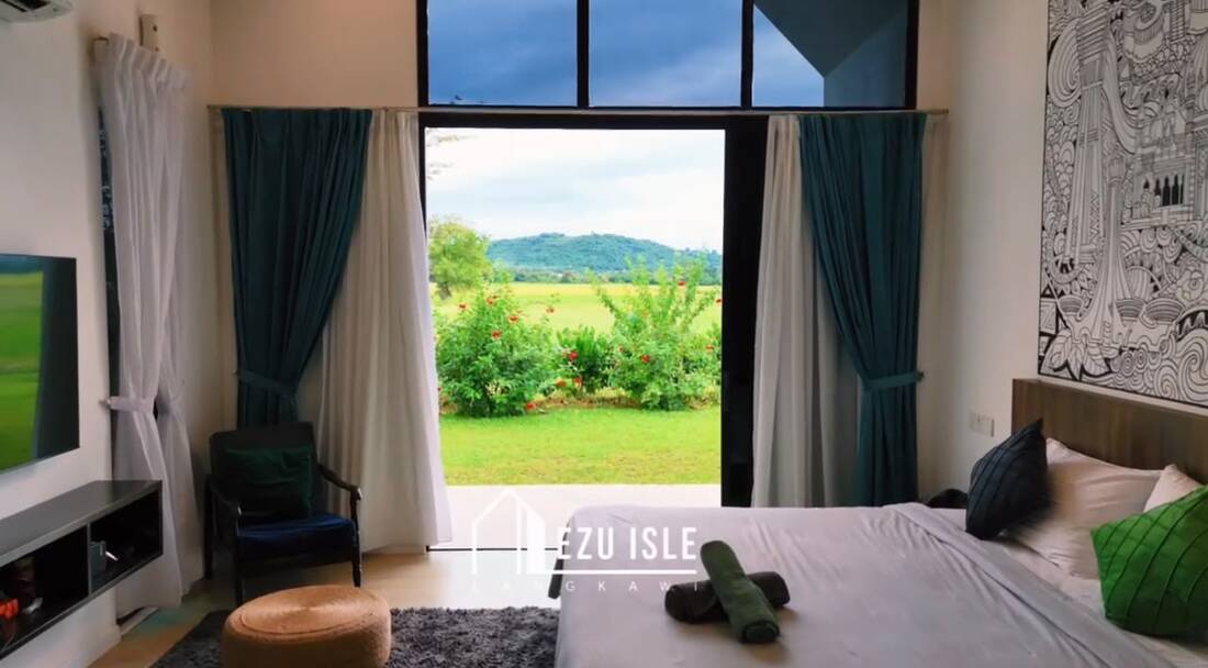 View from a suite bed: doors open onto the lawn, flowering bushes, padi fields and a green hill beyond