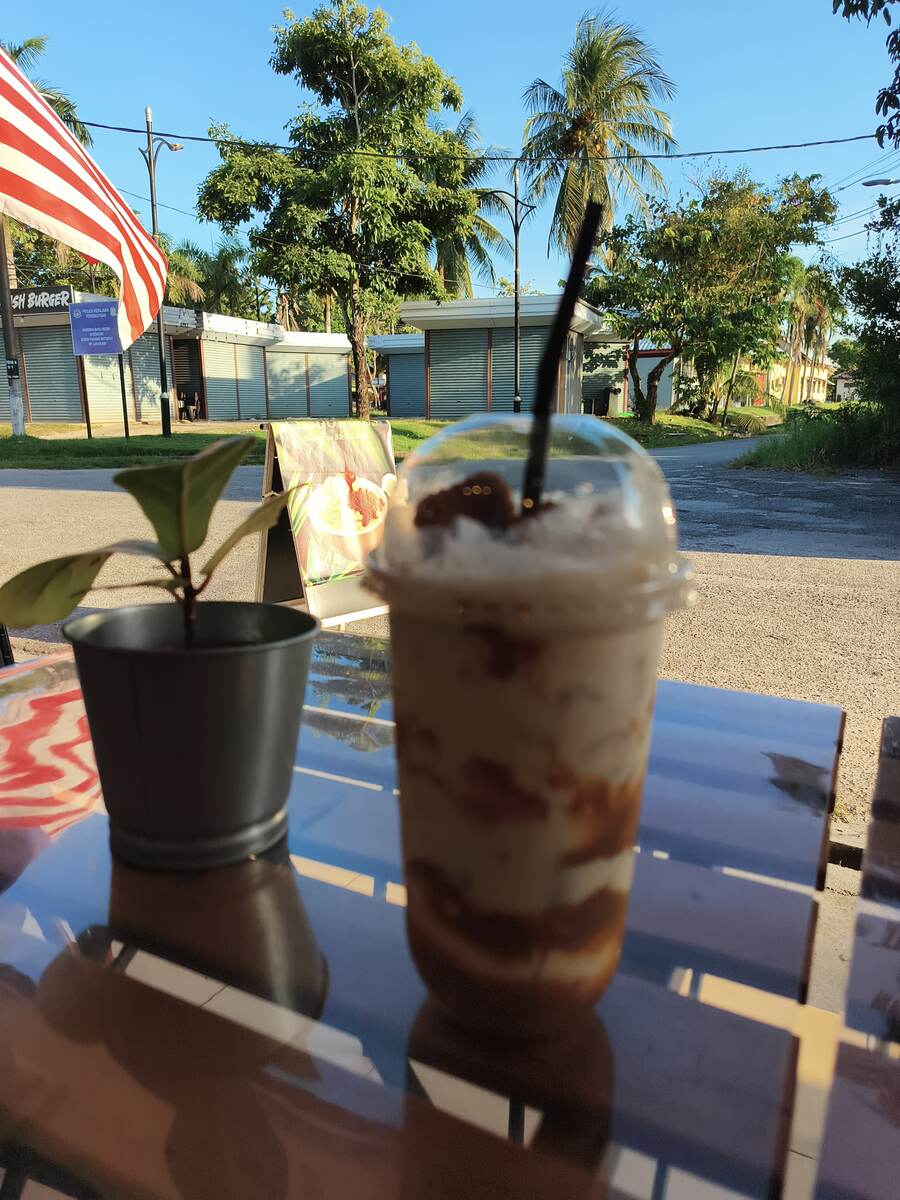 Iced caramel blended drink on an outdoor table with the Malaysian flag and palm trees behind